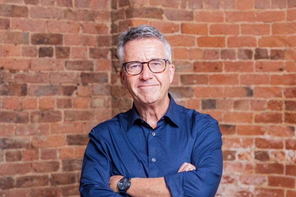 Paul McGee in a blue shirt with arms crossed in front of a brick wall